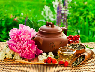 Obraz na płótnie Canvas Chinese tea cerermony in the garden. Clay teapot with glass cup, dry green tea, peony and jasmine flowers, strawberries on a bamboo table.