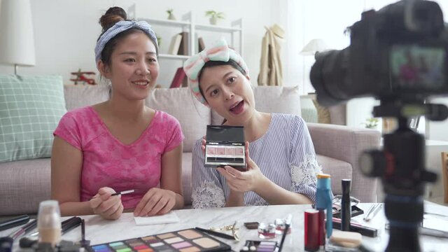two young Asian cute artist women beauty vloggers doing cosmetic makeup tutorial vlog with brushes looking camera on tripod. female recording viral clip share on social media live streaming internet.
