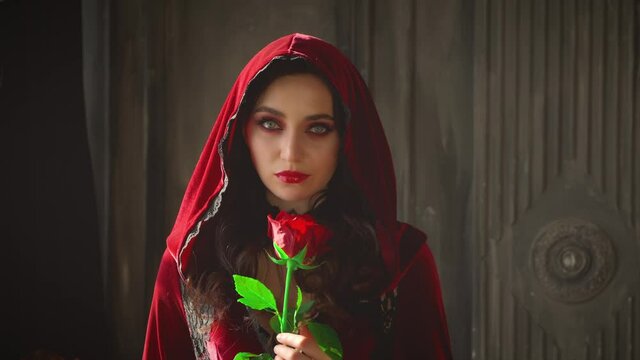 Mysterious beautiful young gothic woman in hood. Closeup woman face with halloween vampire makeup. Sexy fashion velvet vintage red dress. Girl holds rose in hands. Dark room interior medieval castle