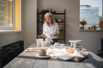 A young european beautiful female potter in a creative process of making ceramics at table in workshop. Hair fluttering in the wind at sunny day at pottery studio.
