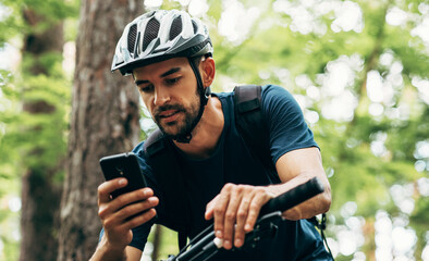 Fototapeta Professional cyclist resting in the forest, holding mobile phone, using online application for searching GPS coordinates while riding the bike. Travel, sport and modern technology concept. obraz