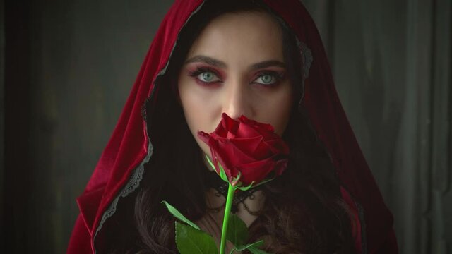 Closeup portrait young mystical vamp woman. Medieval angry witch holding red rose in hands. Vampire sexy vintage gothic dress hood. Art makeup halloween. Gray eyes bright lips. Black necklace