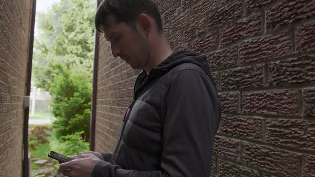 High quality 4k video of a young man who is texting messages on his cellphone standing in a shade of the red bricks wall. Guy hiding between the walls and holding mobile phone in his hands.