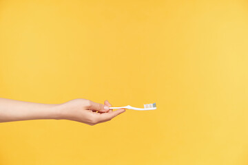 Side view of young female's hand with nude manicure reaching out toothbrush while going to wet it before cleaning teeth, posing over orange background