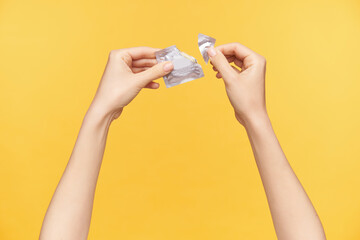 Indoor photo of young woman's hands with nude manicure tearing packaging with condom while having continued date, isolated over orange background