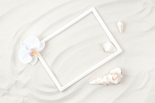Top view of white photo frame on sand background decorated with orchid flowes and seashells. Summer and vacation concept. Flat lay. Copy space.
