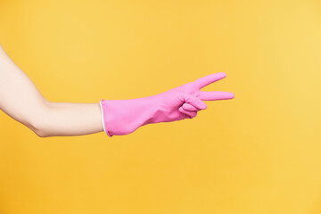 Studio photo of young woman's hand in rubber glove forming victory gesture with two fingers while being isolated over orange background. Signs and gesturing concept