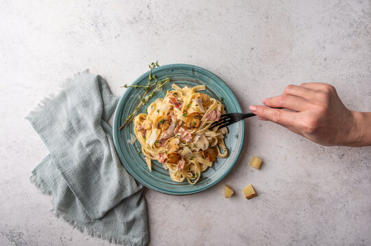 Woman's hand holds a fork with pasta fettuccine with mushrooms, bacon, parmesan and bechamel sauce in plate on a light wooden background, top view