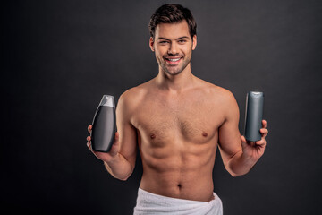 Handsome young man isolated. Portrait of shirtless muscular man is standing on grey background with shampoo in hands. Men care concept.