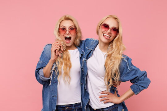 Indoor photo of young lovely blonde twins with wavy hairstyle embracing each othes while laughing and having fun, standing over pink background in casual clothes