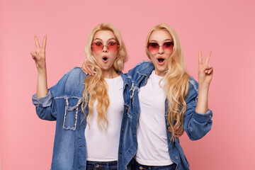 Surprised young attractive white-headed females with wavy hairstyle showing peace gesture and...