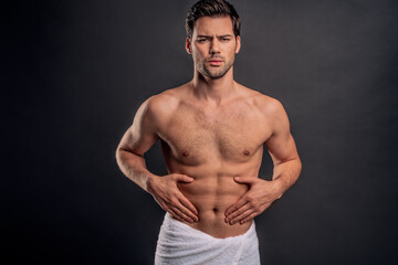 Handsome young bearded man isolated. Cropped image of shirtless muscular man is standing on gray background. Man holding his stomach. Experiencing stomach pain.