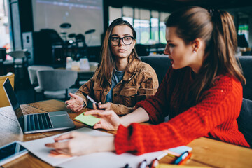 Selective focus on serious hipster girl in spectacles sitting college campus listening carefully female friend, two concentrated women studying together thinking on startup project using technology