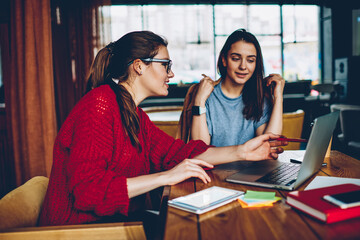 Beautiful hipster girl in spectacles showing information on webpage to her female friend sitting near, two charming women spending time together with laptop device watching videos online indoors