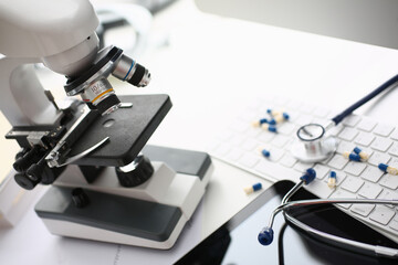 Close up of white table with laboratory optical microscope, pharmaceutical drugs, keyboard and medical device for listening to internal sounds of body