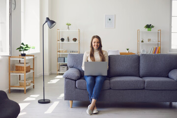 Young beautiful girl uses a laptop while sitting on a sofa in the apartment. Woman works office at...