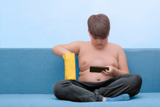  full child sits on a sofa playing a phone and eating snacks. Obesity in children. Copy space.
