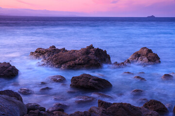Ocean waves rolling over a rocky shore under a purple sunset sky. Long exposure photo with "misty" water