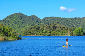 Lake surrounded by forest with a paddle boarding figure on the water. Photographed at Lake Okareka near Rotorua, New Zealand