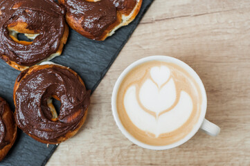Coffee with a drawn heart and milk on a wooden table in a coffee shop. Chocolate donut with scattering on the table next to the coffee.