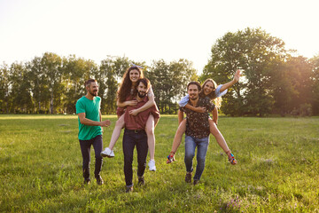 Millennial friends having fun outdoors on summer weekend. Two young guys giving their female friends piggyback ride