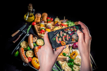 Hands taking photo a grilled sausage and mixed vegetables on a dark background with smartphone