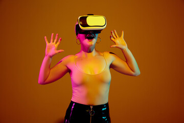 Playing emotional. Young caucasian woman on brown studio background in neon light. Beautiful model with VR-headset. Human emotions, facial expression, sales, ad concept. Freak's culture. Copyspace.