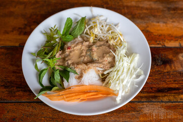 Rice Noodle eaten with curry and fresh vegetable in a white plate on a wooden table background. Fermented Rice Flour Noodles/kanom jeen are fresh rice noodles in Thai cuisine which are made from rice.