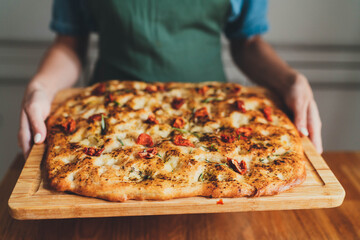 Closeup of woman in green apron holding fresh baked homemade Italian focaccia with rosemary and sun-dried tomatoes on wooden board, Homemade Food Concept