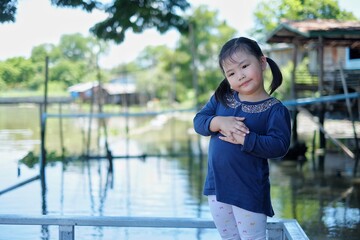 A cute young Asian girl standing on a wooden pier by a river, patiently waiting for her boat ride, smiling.