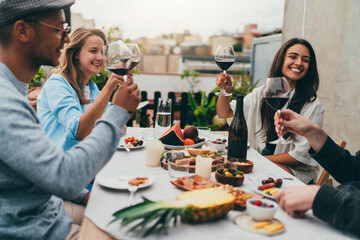 Multi ethnic group of happy friends having dinner party on rooftop sitting at festive table laughing and joking while raising toasts with red wine, Friendship Dining Celebration Hanging out Concept