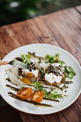 Photo of white plate with parsley, poached eggs, tomatoes, seeds and sauce on wooden table in blurred background