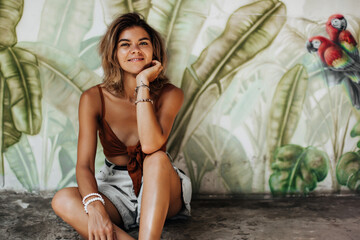 Happy young woman in brown top and denim shorts widely smiles and sits on floor. Pretty tanned girl in summer outfit poses on background of palm leafs