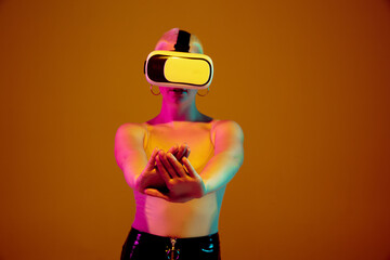 Touching, pointing on copyspace. Young caucasian woman on brown studio background in neon light. Beautiful model with VR-headset. Human emotions, facial expression, sales, ad concept. Freak's culture.