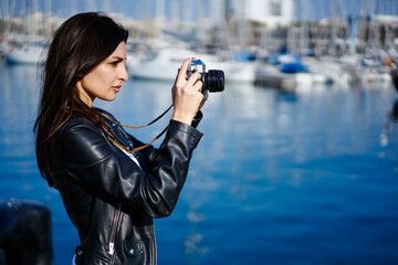 Concentrated brunette photographer making picture of urban setting walking on sunny day in sea port, charming tourist taking photo via vintage camera standing near blurred promotional background