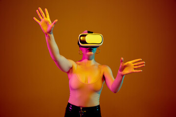 Touching, pointing on copyspace. Young caucasian woman on brown studio background in neon light. Beautiful model with VR-headset. Human emotions, facial expression, sales, ad concept. Freak's culture.