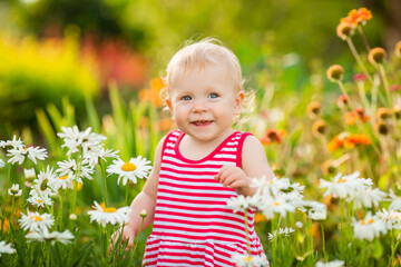 Baby girl smiles in summer dress walking outside among daisies in garden