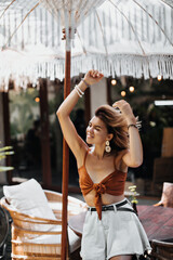 Blonde tanned woman with big white earrings and dressed in brown top and denim shorts ruffles hair....