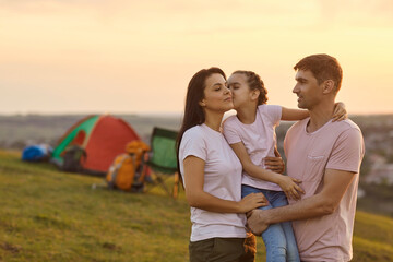 Young man holding his daughter and kissing his beautiful wife at camp in mountains.