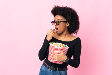 Young African American woman isolated on pink background with 3d glasses and holding a big bucket...