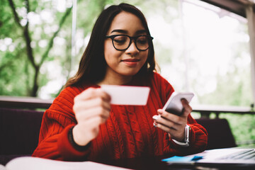 Pensive young woman in eyeglasses dialing number on mobile from visit card for contact service...