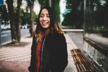 Obraz premium Half-length portrait of cheerful asian woman enjoying free time outdoors standing on bus stop, happy hipster girl in casual outfit laughing looking at camera walking on city street on weekend