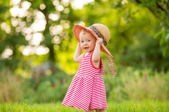 Cute little girl in a red dress and hat walks in summer barefoot on the green grass, space for text