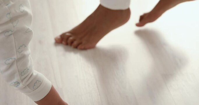 Close up young barefoot african american mother holding hands of little toddler kid son, teaching walking on warm floor. Small infant child daughter making first steps with caring mommy at home.