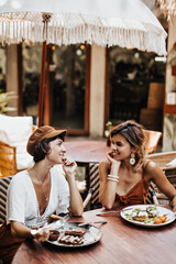 Two girl in summer stylish outfits talking and eating delicious food in street cafe. Brunette woman in white tee and tanned blonde lady in brown bra chilling out and enjoys waffle