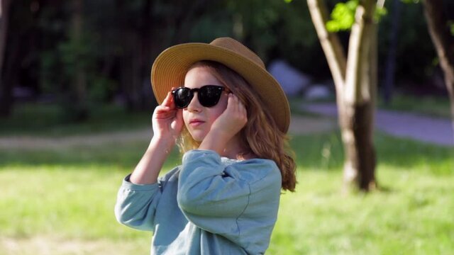 Little girl in a straw hat puts on black sunglasses and smiles at camera
