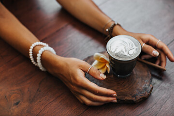 Fototapeta na wymiar Photo of female tanned hands holding cup of coffee with milk. Snapshot of glass of latte, yellow flower on wooden board