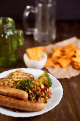 Grilled vegetables with sausage and Mexican corn chips nachos lie on a wooden table.