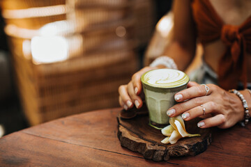 Female hands with silver rings holds glass of matcha latte. Woman in brown bra poses with cup of green tea with milk, yellow flower on wooden table