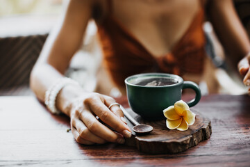 Woman in brown bra touches peace of wood. Photo of porcelain blue cup of coffee or tea and yellow tropical flower